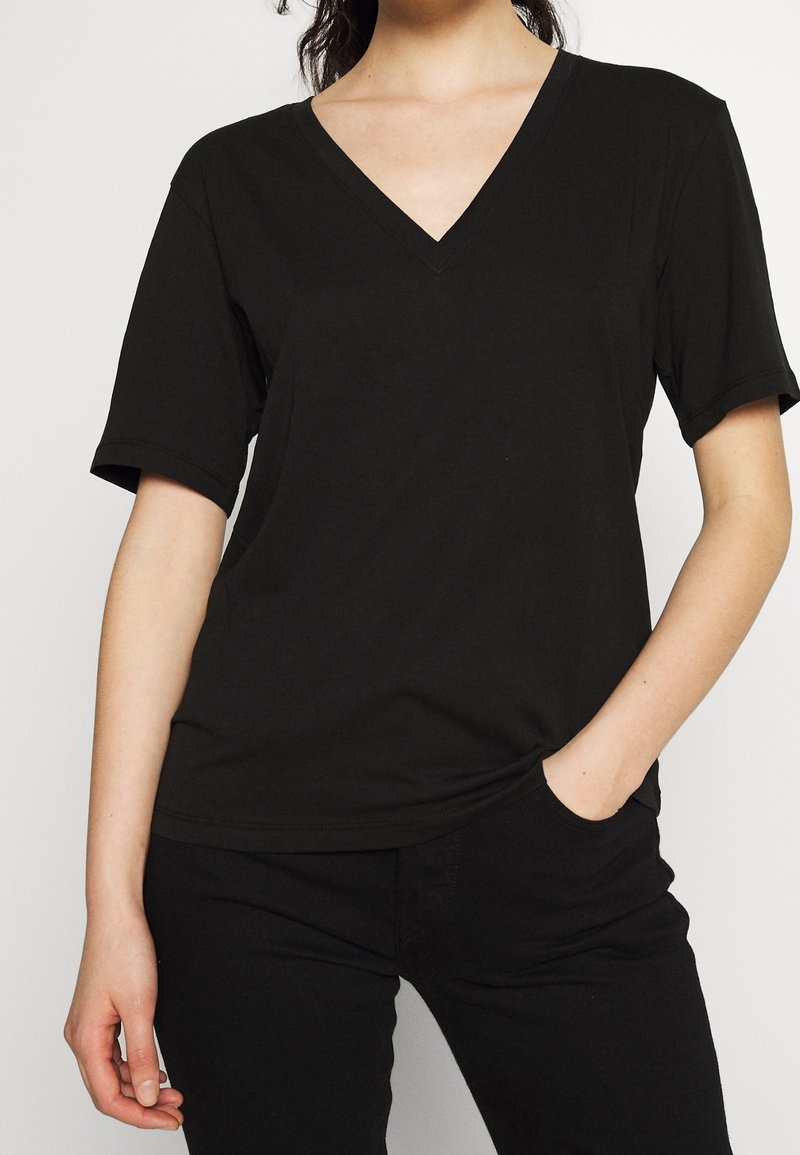 Personne portant un t-shirt noir uni à col en V et manches courtes ainsi qu'un pantalon noir, une main dans la poche, sur fond blanc.