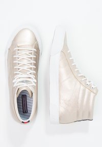 Zapatillas altas metálicas doradas con cordones blancos y suelas blancas de goma. Parte superior de cuero texturizado con interior de tela a rayas y detalle del logo.