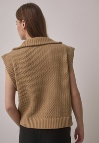 Knit, sleeveless beige sweater with a large collar, featuring open weave texture and ribbed hem, worn over a dark outfit.