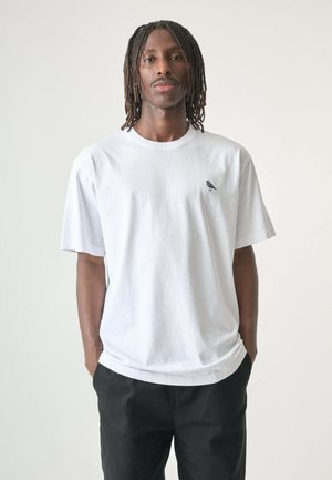 White cotton t-shirt with a round neckline, short sleeves, and a small black bird logo on the left chest. Worn with black pants.