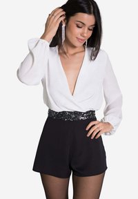 Body à manches longues blanc avec un décolleté en V profond, associé à un short noir. La taille présente un détail en sequins noirs. Modèle avec des cheveux foncés.