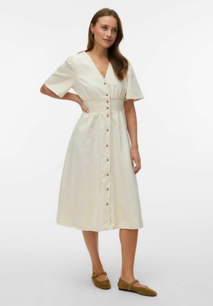 Kvinde står med hånden på hoften iført en cremefarvet, knappet midi-kjole med korte ærmer og brune flade sko.