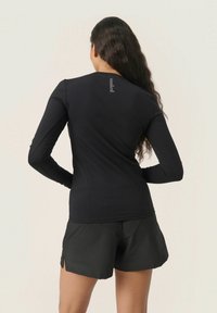 Maglietta nera a maniche lunghe con design aderente, realizzata in tessuto liscio, con un logo discreto sul retro e maniche lunghe, abbinata a pantaloni corti neri.