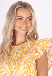 Bluză cu mâneci scurte, în alb și galben cu un model floral, având mâneci volănașe și un decolteu rotund. Textură de stofă netedă.