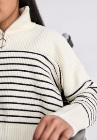 Maglione crema a righe con colletto alto, dotato di maniche lunghe e texture a costine. Le righe sono nere su una base chiara.