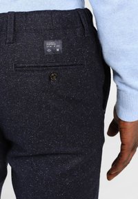 Marinblå byxor med en texturerad tyg, som har en bakficka med knappstängning och en märkesetikett nära midjan.