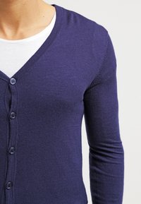 Marinblå stickad kofta med V-hals, långa ärmar och knapplist med fem rundade knappar. Mjuk, slät struktur.