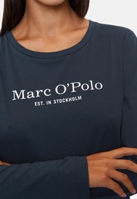 T-paita pitkällä hihalla, tummansininen, puuvillasta valmistettu, jossa pyöreä kaula-aukko ja valkoinen teksti: "Marc O’Polo EST. IN STOCKHOLM" etupuolella.