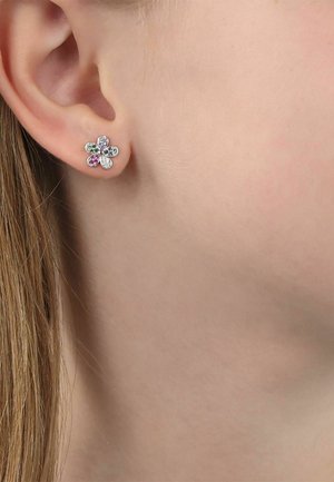 Pendiente de botón con forma de flor y pequeñas piedras preciosas multicolores, llevado en el lóbulo de una persona de piel clara y cabello castaño.