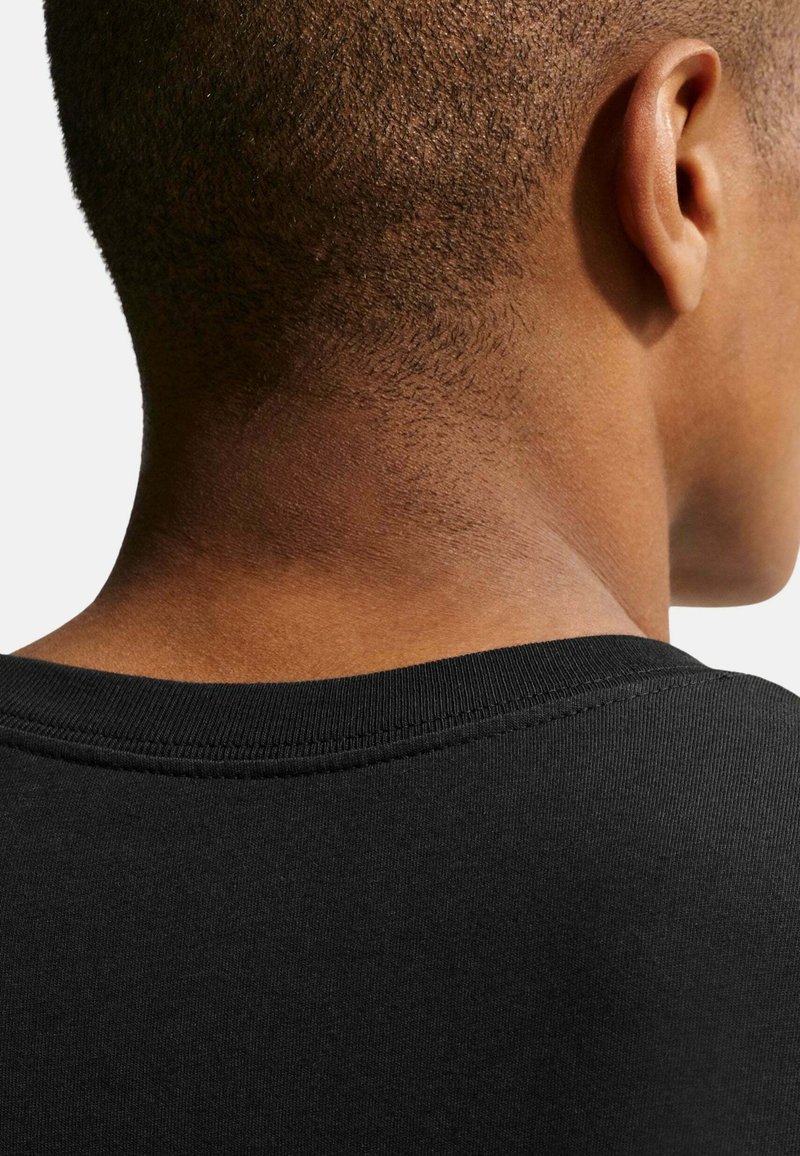 Parte trasera de la cabeza de una persona con cabello corto, oreja visible, vistiendo una camiseta negra de cuello redondo sobre un fondo claro y sencillo.