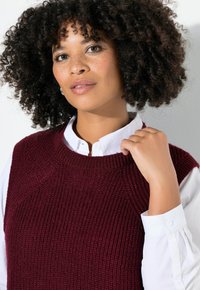 Chaleco de punto burdeos con textura acanalada, escote redondo, llevado sobre una camisa blanca de botones, mostrando un estilo de capas casual.