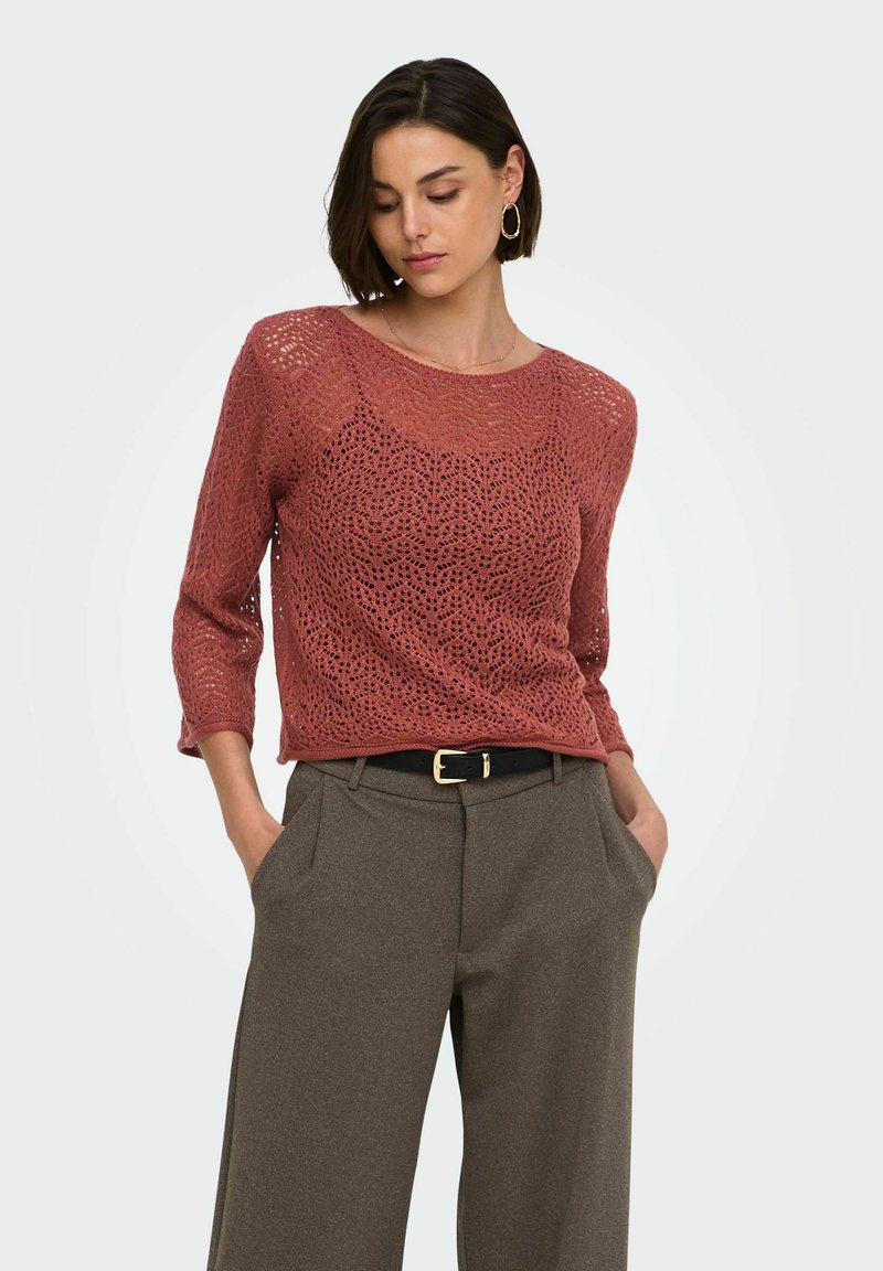 Femme portant un pull à mailles ajourées couleur rouille avec des manches trois-quarts et un pantalon marron taille haute avec ceinture noire.