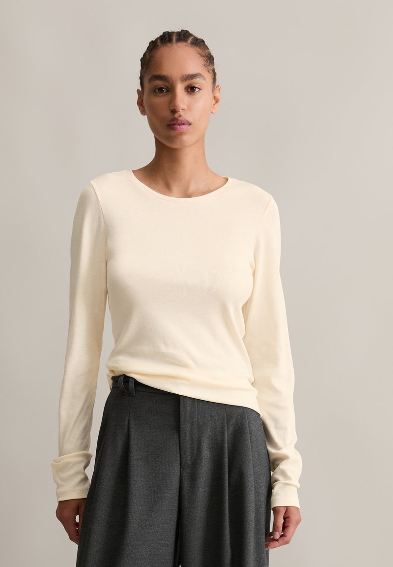 Helles cremefarbenes Langarmoberteil aus einem weichen Strickstoff, mit Rundhalsausschnitt und enger Passform, kombiniert mit dunkelgrauen High-Waist-Hosen.