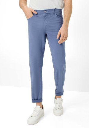 Uomo con pantaloni azzurri risvoltati e sneakers bianche, in piedi con una mano in tasca contro uno sfondo bianco.