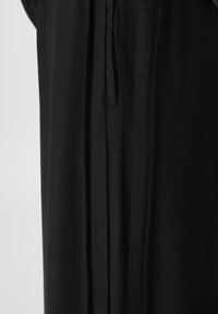 Μαύρο ένδυμα με υφή σε σχέδιο βάφλας. Διαθέτει εμφανείς ραφές και κάθετη σχισμή, που αναδεικνύει ένα απαλό, ρέον ύφασμα.