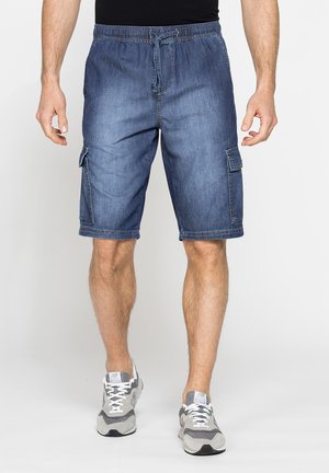 Uomo con pantaloncini cargo in denim blu con coulisse in vita e scarpe da ginnastica grigie, in piedi davanti a uno sfondo semplice.