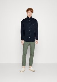 Marineblauwe overhemd met knopen en een zak, groene taps toelopende broek en beige instap schoenen. Effen textuur, casual ontwerp, geen zichtbare patronen.