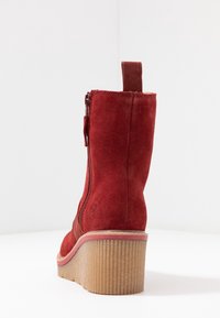 Red suede ankle boot with a zipper closure, rounded heel, and textured rubber sole. Features a pull tab on the back for easy wear.