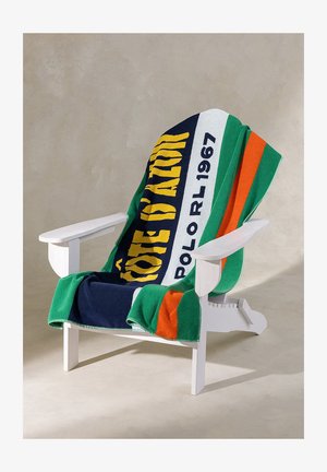 Witte houten stoel met een oversized, kleurrijke handdoek eroverheen gedrapeerd, met groene, marineblauwe, oranje en gele afbeeldingen.