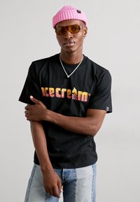 Sort kortærmet t-shirt med "icecream" logo i multicolor. Modellen har lyseblå jeans og en lyserød strikket hue med solbriller på.