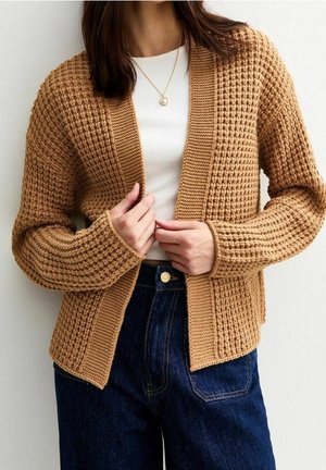 Vrouw die een bruine gebreide cardigan met textuur draagt over een wit shirt en een blauwe spijkerbroek, haar cardigan met beide handen verstelend.