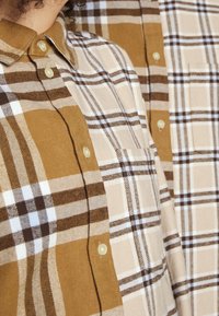 Twee geruite overhemden tentoongesteld, een in bruintinten met witte strepen, een in beige met bruine strepen. Beide hebben knoopsluitingen en borstzakken.