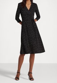 Svart omlottklänning med trekvartslånga ärmar, med ett spritt mönster i vitt och guld. A-linjeformad silhuett med ett midjeband.