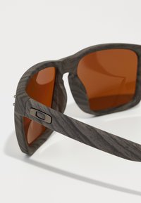 Gafas de sol con patrón gris y lentes naranjas, que presentan un marco texturizado. Un detalle metálico en el costado añade un toque de durabilidad al diseño.