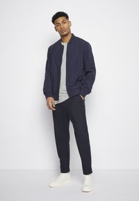 Casaco bomber azul-marinho com punhos canelados, t-shirt cinza, calças escuras e sapatilhas brancas de cano alto, de pé contra um fundo branco.