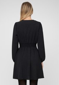 Vestido negro con mangas largas abullonadas, cintura fruncida y falda acampanada. Tela suave, diseño sencillo, con largo hasta el muslo.