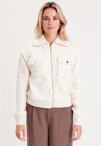 Chaqueta de cremallera color crema con una tela suave y texturizada en la mitad superior, que presenta dos bolsillos frontales y puños y dobladillo acanalados.