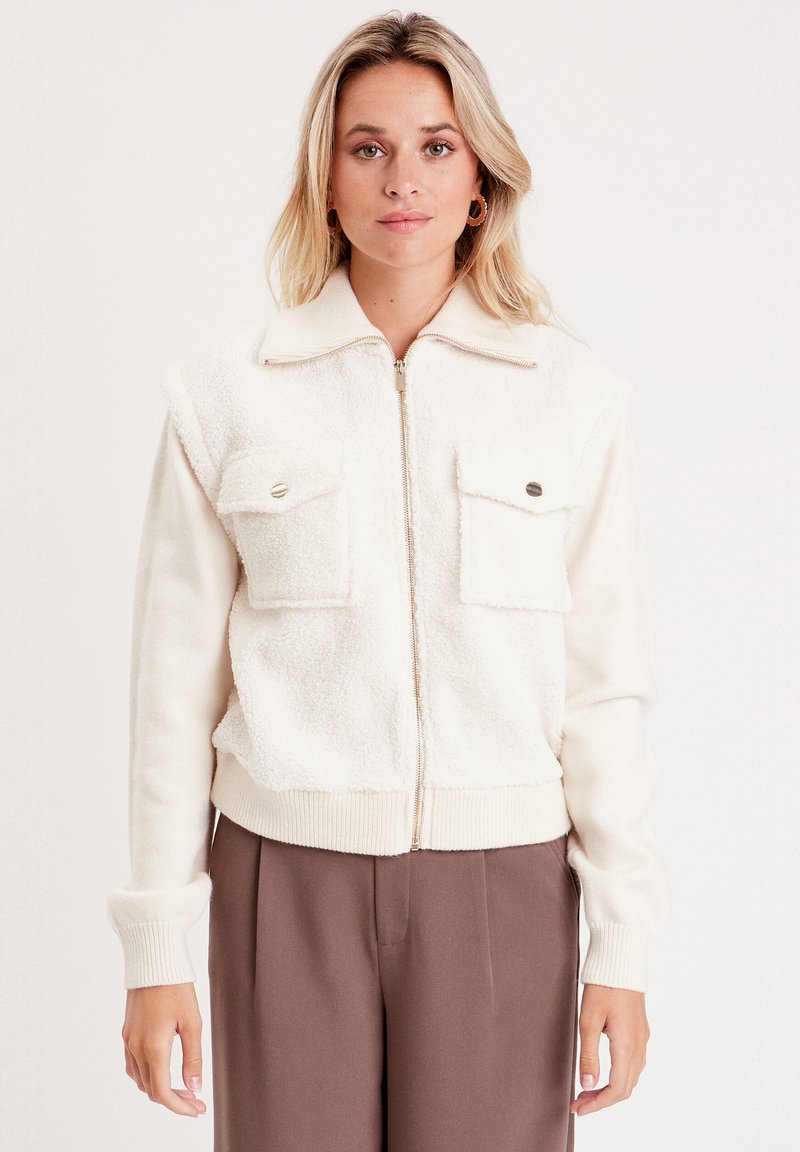 Chaqueta de cremallera color crema con una tela suave y texturizada en la mitad superior, que presenta dos bolsillos frontales y puños y dobladillo acanalados.