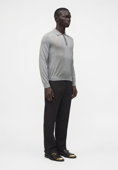 Homme debout portant un polo gris clair à manches longues, un pantalon noir et des chaussures noires avec des bouts dorés, sur un fond blanc uni.