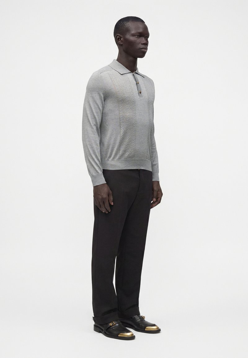 Homme debout portant un polo gris clair à manches longues, un pantalon noir et des chaussures noires avec des bouts dorés, sur un fond blanc uni.