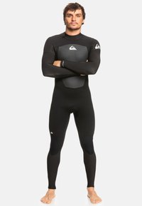 Man die blootsvoets staat met zijn armen gekruist, gekleed in een zwart wetsuit dat het hele lichaam bedekt, met grijze patches op de borst en knieën.