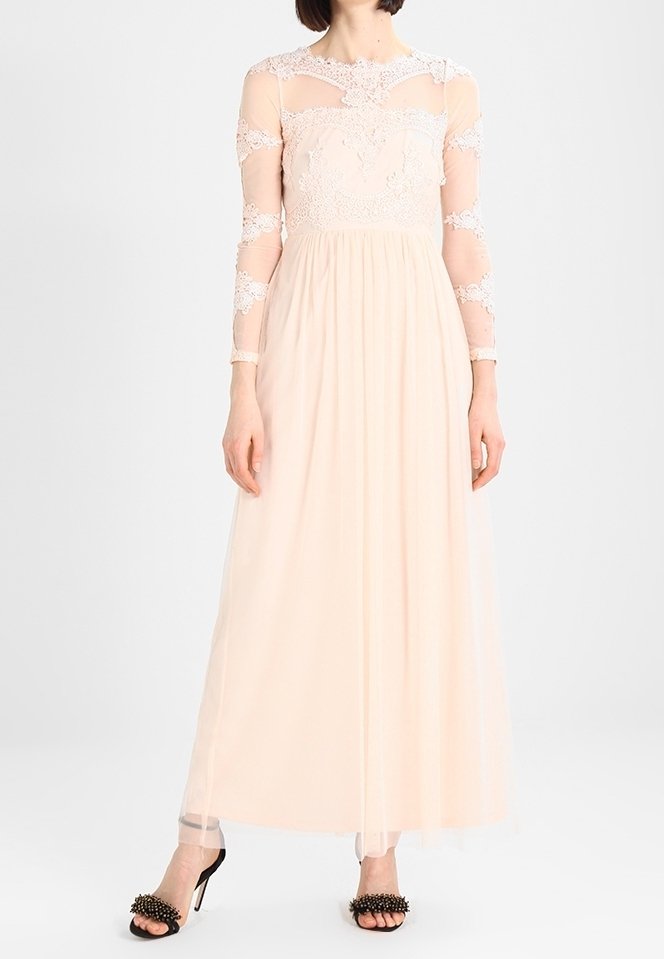 Femme portant une longue robe rose poudré avec manches et corsage en dentelle, associée à des sandales noires à talons hauts avec des pointes décoratives.