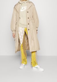 Beige trenchcoat med huva, lager på lager över en ljusgrön huvtröja. Utsmyckad med gula virkade byxor och vita sneakers med gröna detaljer.