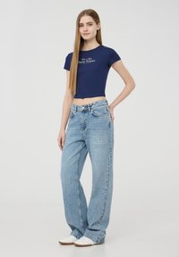 Maglietta corta blu navy con logo stampato, abbinata a jeans azzurri a gamba larga. Modello indossa sneakers bianche, in piedi su uno sfondo neutro.
