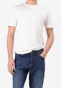 Man i en enkel vit kortärmad t-shirt och mörkblå denimjeans, med en hand i fickan framför en vit bakgrund.
