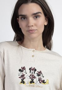 Camisa de manga larga color beige claro con tres gráficas de Minnie Mouse en vestidos de lunares rojos. Presenta un escote redondo y dos tapetas de botones.