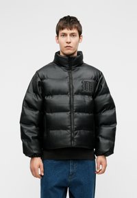 Jovem vestido com uma jaqueta de couro acolchoada preta, com gola alta e logótipo bordado, combinada com jeans azuis, em pé contra um fundo simples.