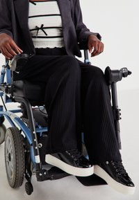 Μαύρο ριγωτό παντελόνι σε φαρδιά γραμμή, παπούτσια από παγωμένο δέρμα σε μαύρο χρώμα με λευκές σόλες, και μία υφασμάτινη λευκή μπλούζα με μαύρες ρίγες και φάσες, φορεμένα σε αναπηρική καρέκλα.