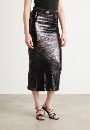 Femme portant une jupe midi noire à sequins, un top noir sans manches et des sandales noires à talons hauts à brides, debout sur un sol gris.