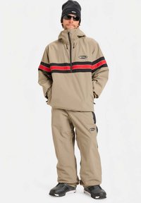 Homem vestido com um fato de esqui bege com riscas pretas e vermelhas, gorro preto, óculos de sol e botas de neve pretas, em pé com as mãos nos bolsos.