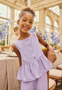 Lila ujjatlan peplum felső három gombos díszítéssel, puha textúrával, hozzáillő lila nadrággal párosítva; a háttér virágos dekorációval díszített.