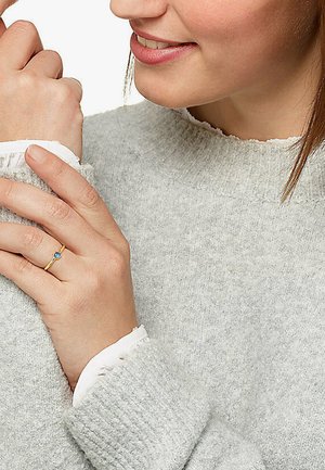 Gold ring with a blue gemstone, worn on a hand. The sweater is gray with a textured knit and white accent at the cuffs.