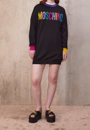Osoba ubrana w czarną sukienkę dresową Moschino z wielokolorowym logo, różowo-żółtymi mankietami oraz czarne sandały na platformie ze złotymi detalami.