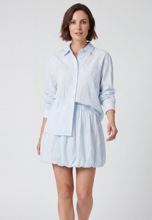 Donna con capelli corti e castani che indossa una camicia azzurro chiaro a maniche lunghe con bottoni e una gonna corta abbinata su uno sfondo chiaro uniforme.
