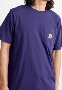 T-shirt di cotone viola con scollo rotondo, maniche corte e tasca sul petto con una piccola etichetta arancione. Texture liscia.