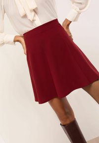 Naine, kes kannab valget pluusi, mis on sisse pistetud kõrge vöökohaga, lehvikukujulisse burgundia seelisse, koos põlveni ulatuvate pruuni värvi saapadega, neutraalse taustaga.